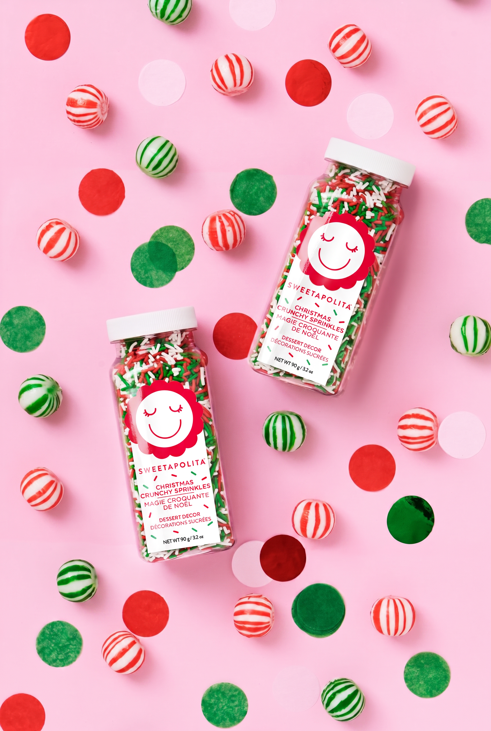 Two jars of Sweetapolita holiday sprinkles filled with red, green, and white jimmies, arranged on a pink background with scattered red, green, and striped candy pieces.