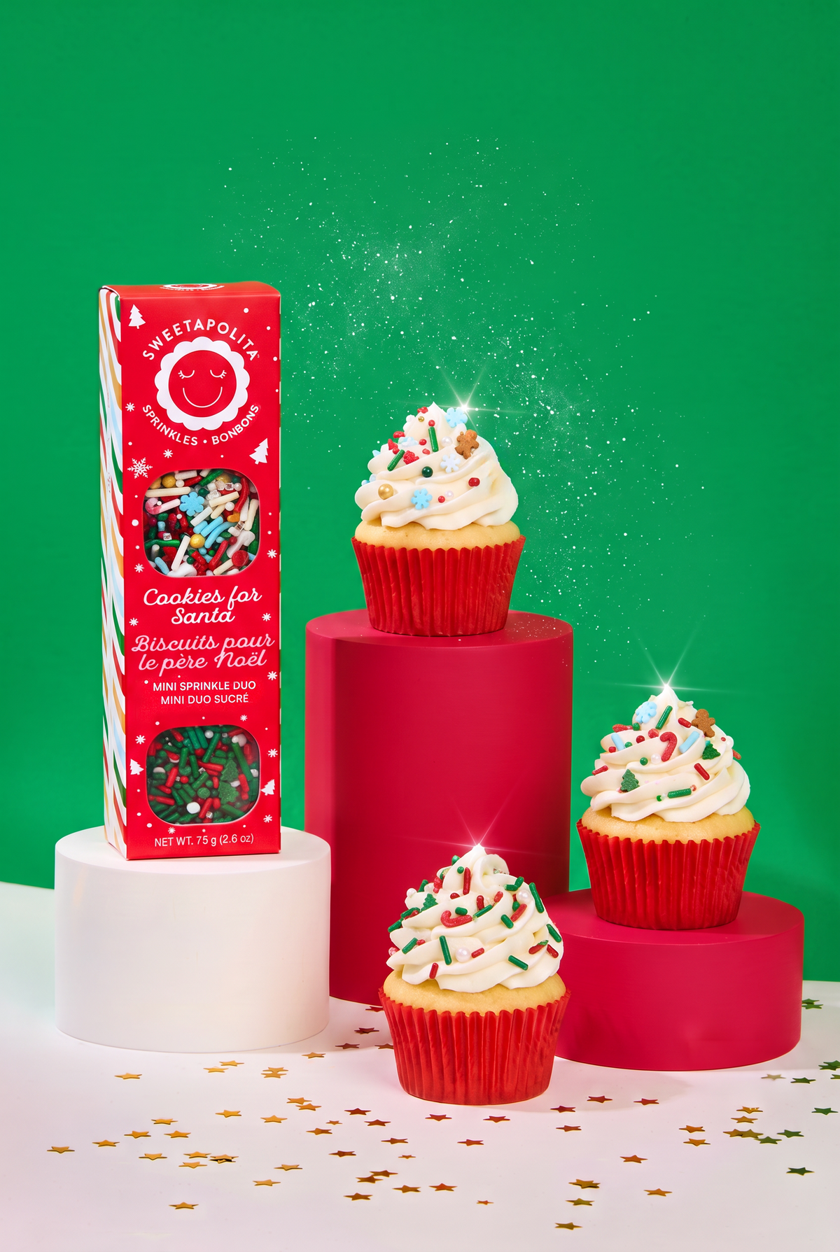 Sweet Treats 'Cookies for Santa' mini sprinkles in a red box, styled with three decorated cupcakes on red and white pedestals against a bright green background.