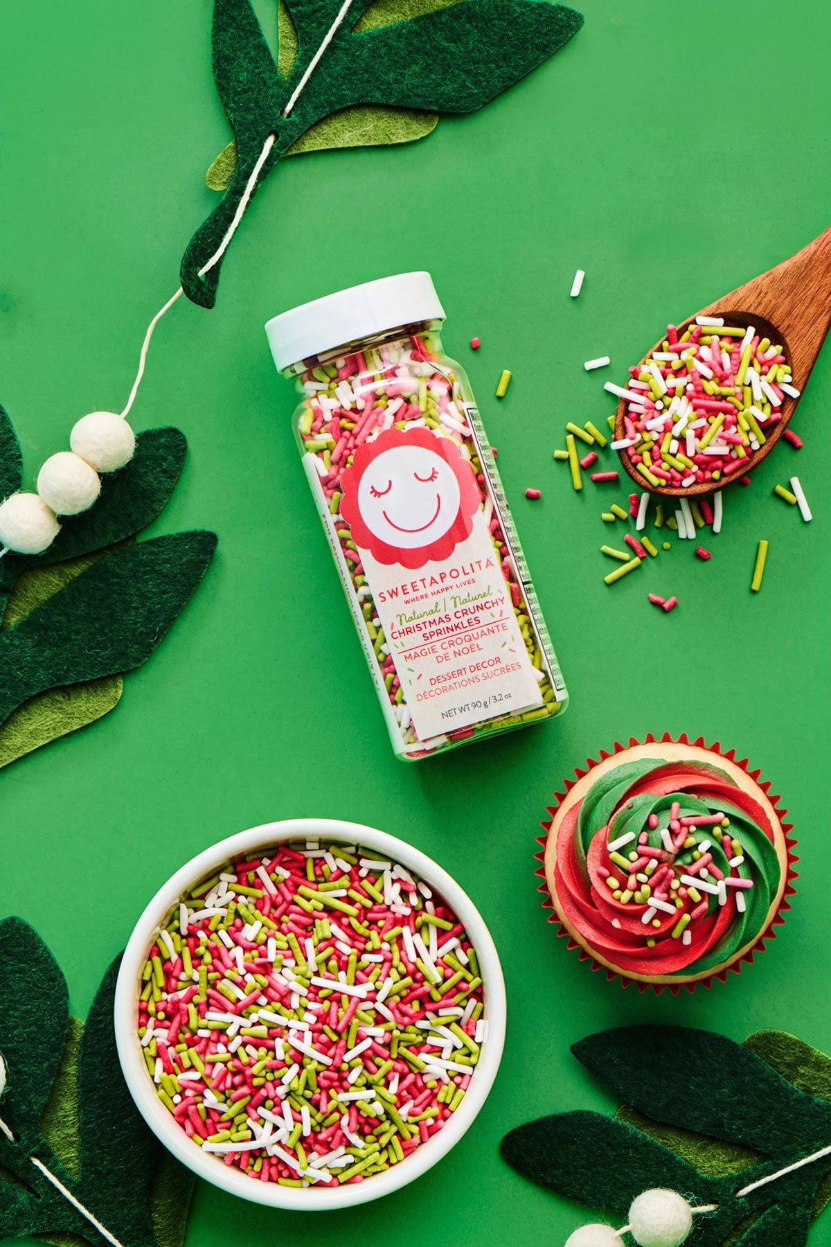 A bottle of Sweetapolita sprinkles displayed on a bright green background with styled props including a white bowl, a wooden spoon, a decorated cupcake, and green felt leaves. The sprinkles are red, green, and white.