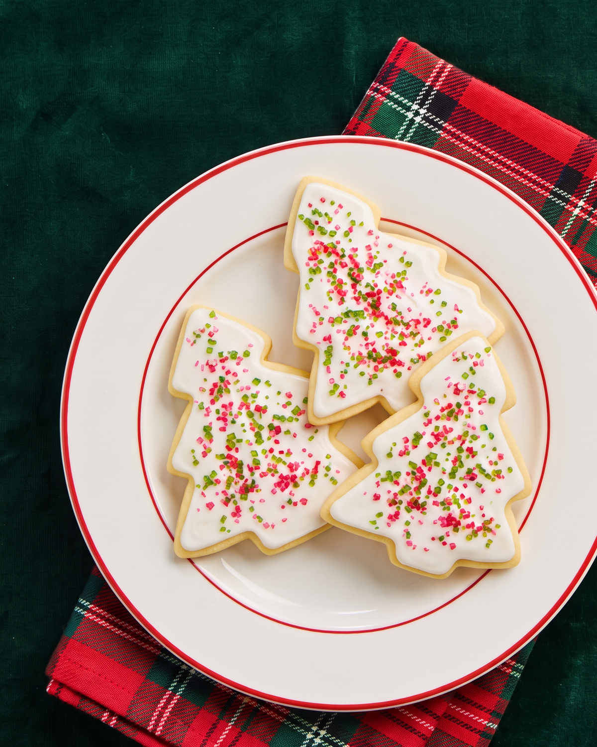 Three Christmas tree-shaped sugar cookies with white icing and red and green sugar, served on a white plate with red trim over a red plaid napkin on a dark green background.