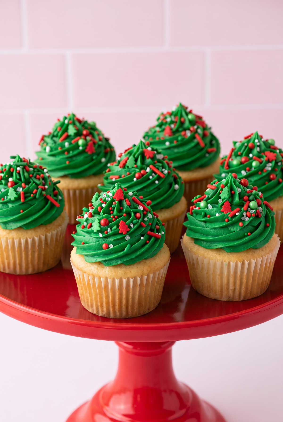 A group of frosted cupcakes with bright green icing and red and green sprinkles, displayed on a red cake stand against a pale pink tiled background.