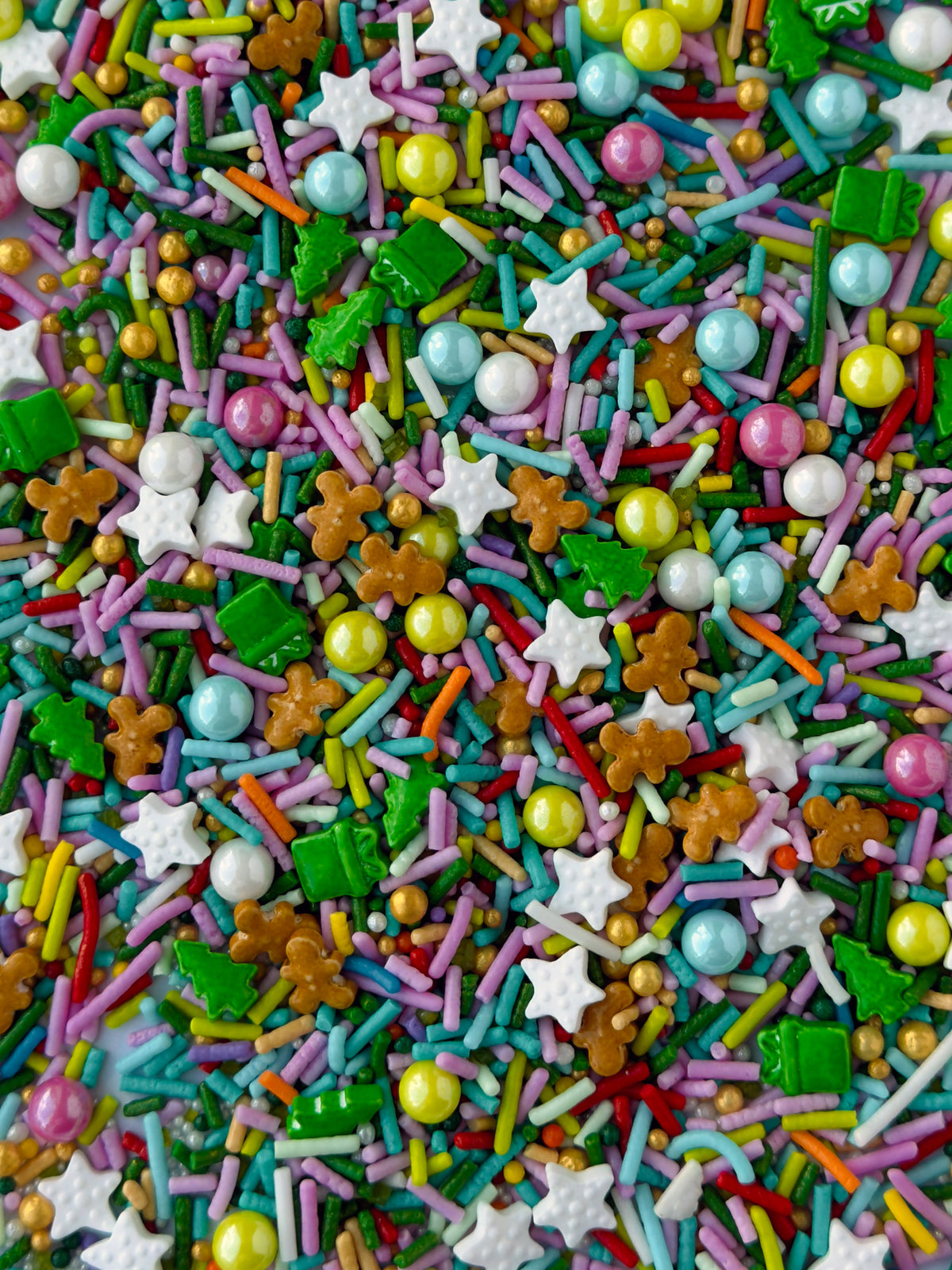 A close-up, top-down view of a colorful sprinkle mix containing white stars, green candy presents, gingerbread men, pastel round beads, and multicolored sprinkles.