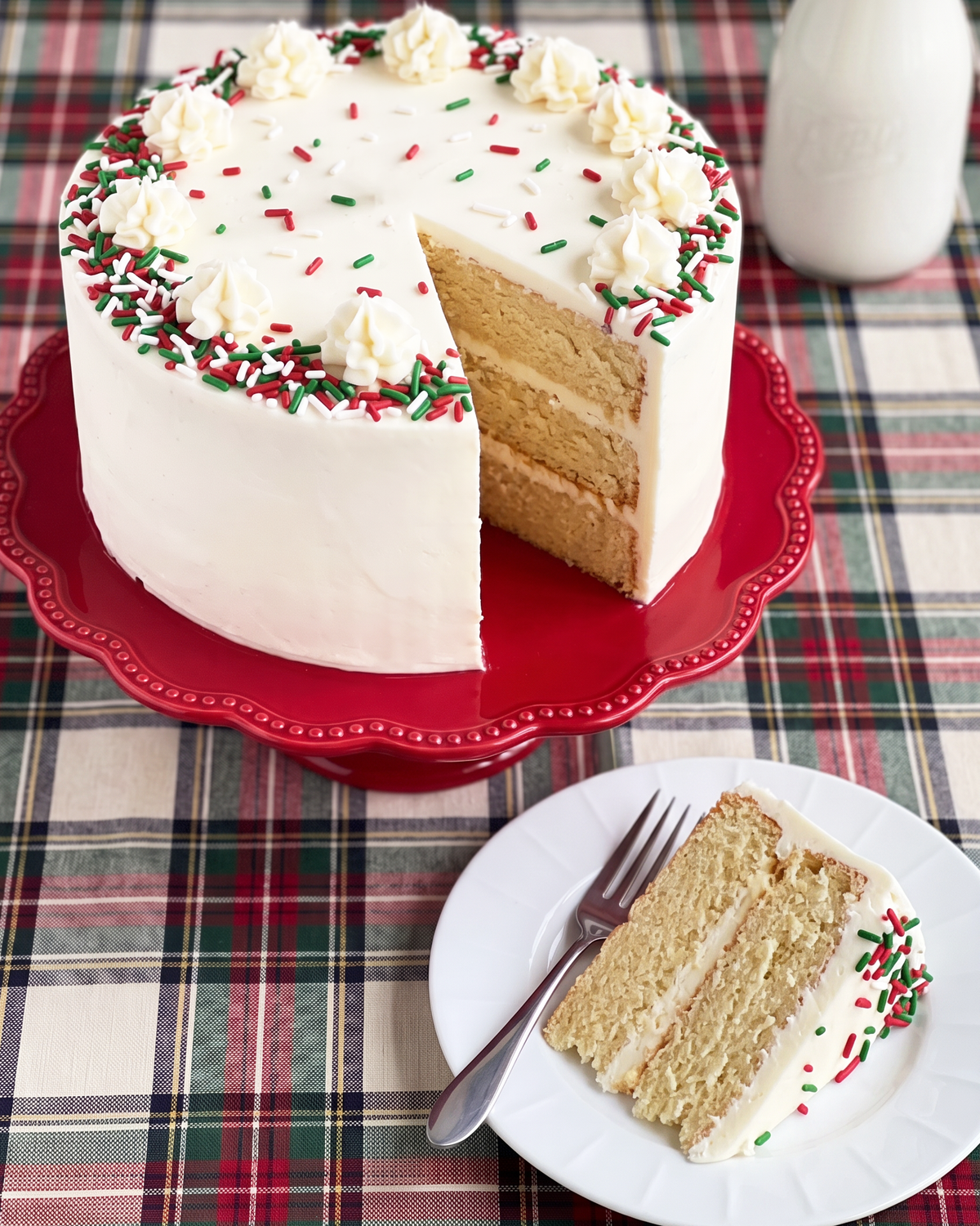 A frosted three-layer cake with white icing, red and green sprinkles, and a slice cut out, displayed on a red cake stand on a plaid tablecloth. A plate with a fork holds a slice in the foreground, and a white pitcher is blurred in the background.