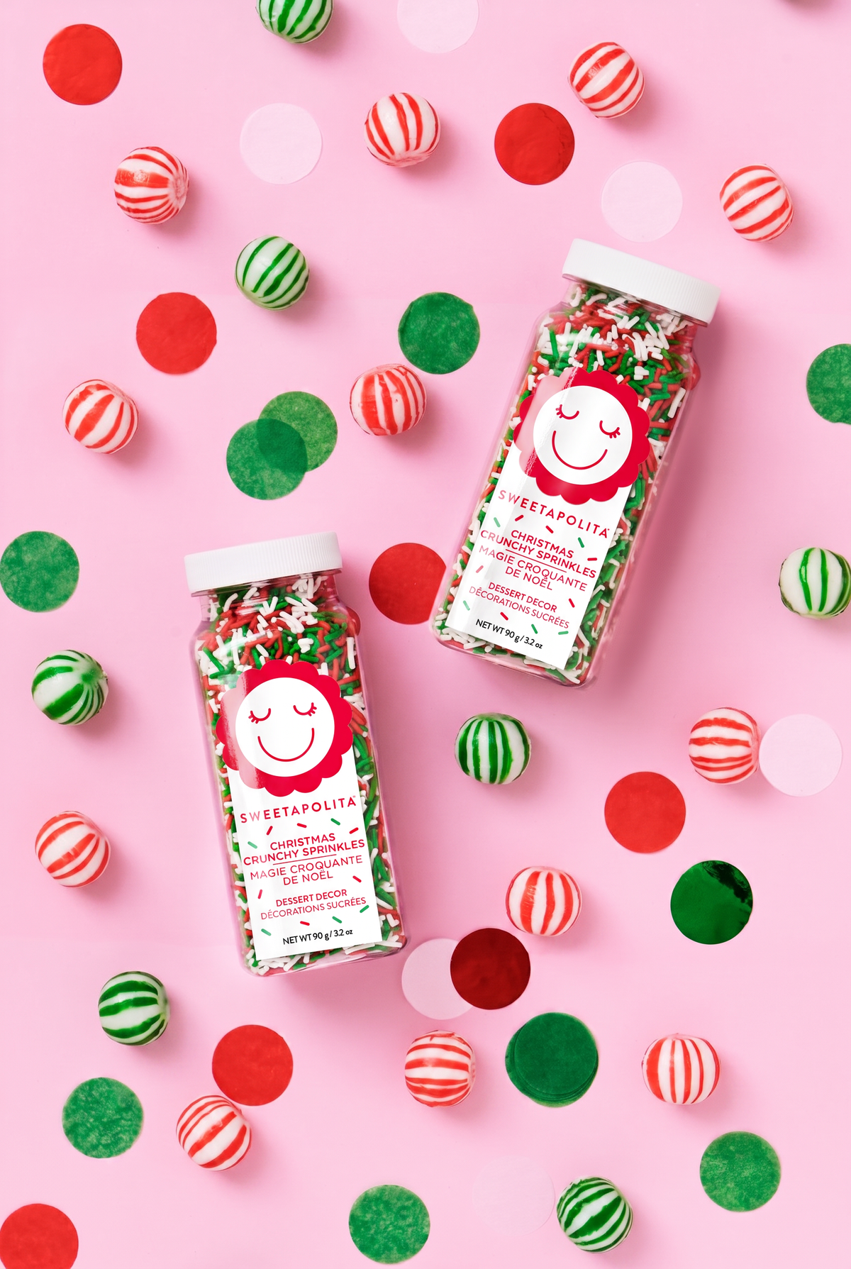 Two jars of Sweetapolita holiday sprinkles filled with red, green, and white jimmies, arranged on a pink background with scattered red, green, and striped candy pieces.