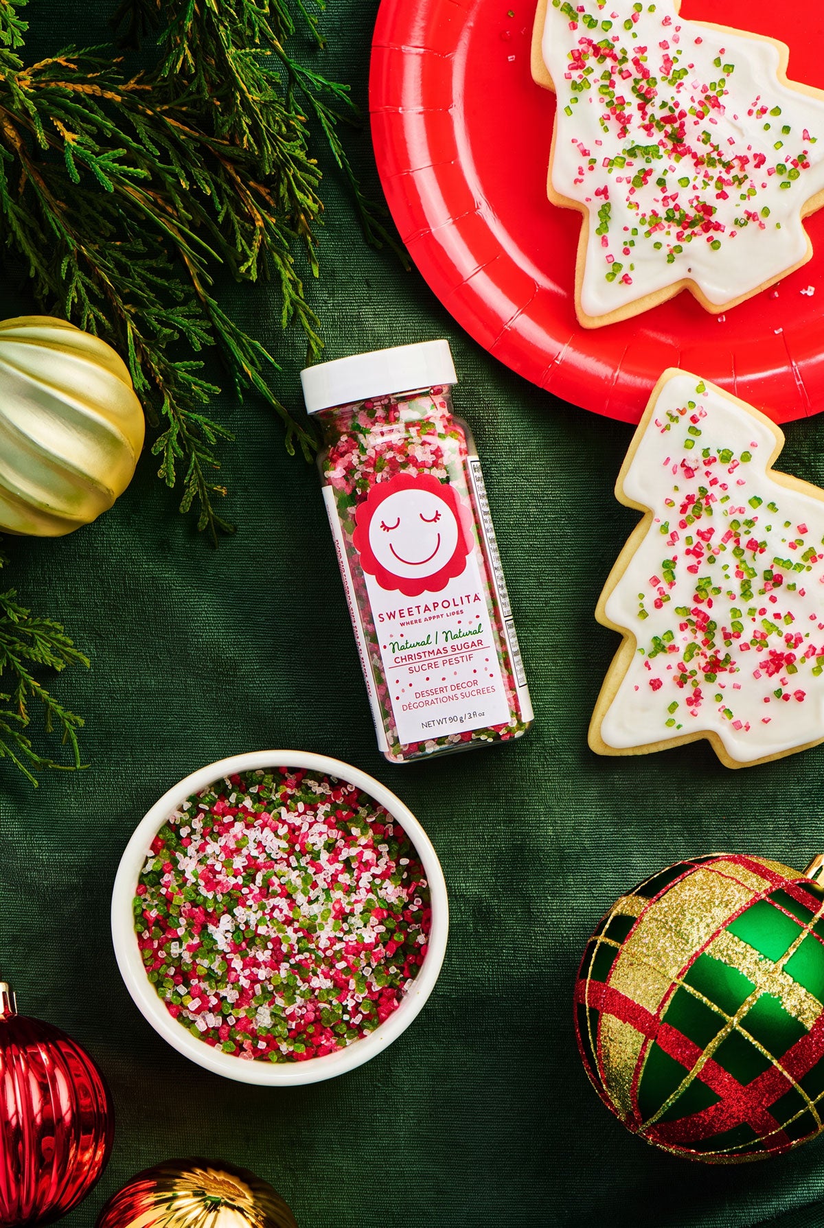 A bottle of Christmas Sugar surrounded by holiday decorations, including a bowl of sprinkles, decorated Christmas tree cookies on red plates, and ornaments on a dark green background.