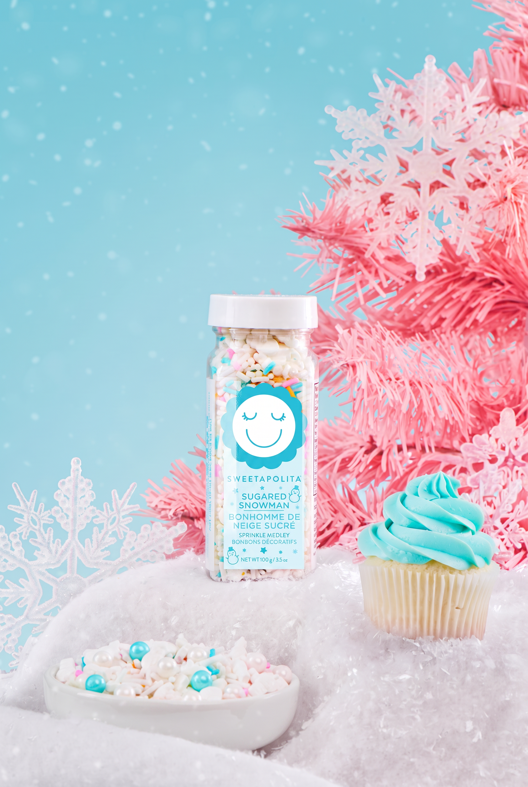 A bottle of Sweetapolita Snowman sprinkles on a white snowy surface next to a cupcake with blue frosting and a bowl of sprinkles, with pink snowflake decorations against a light blue background.