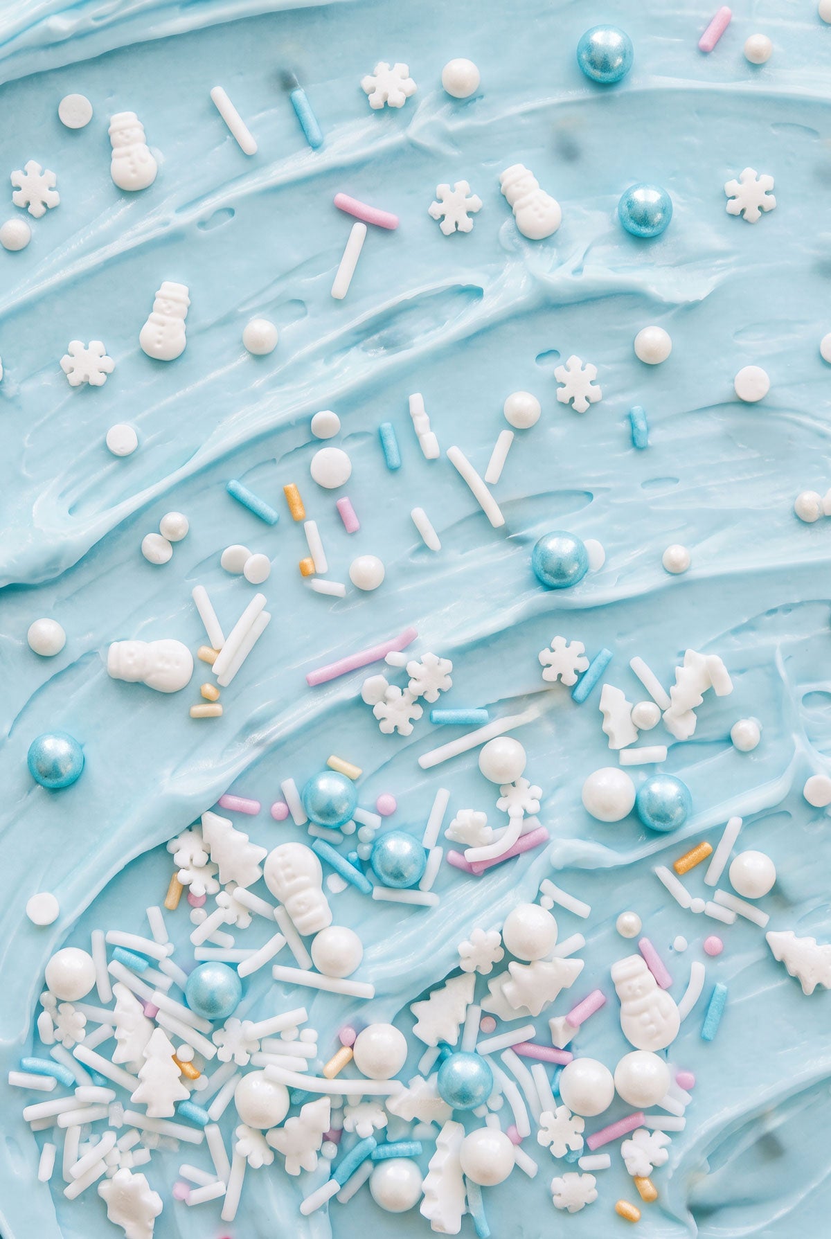 A close-up flat lay of light blue frosting with scattered decorative sprinkles, including white snowflakes, white snowman candies, white tree-shaped pieces, blue pearl-like beads, and pastel pink, blue, and white sprinkles.