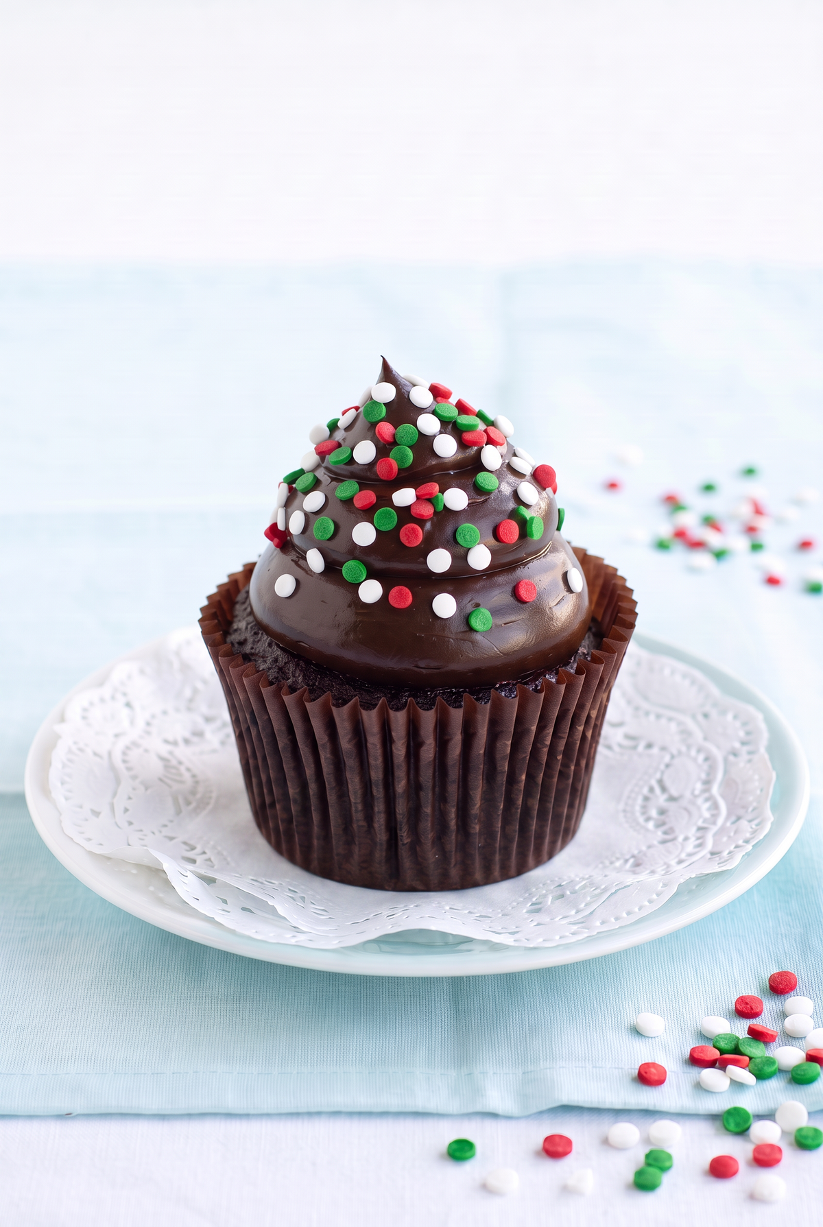 A chocolate cupcake with dark chocolate frosting and red, white, and green sprinkles, presented on a white plate with a lace doily on a light blue surface.