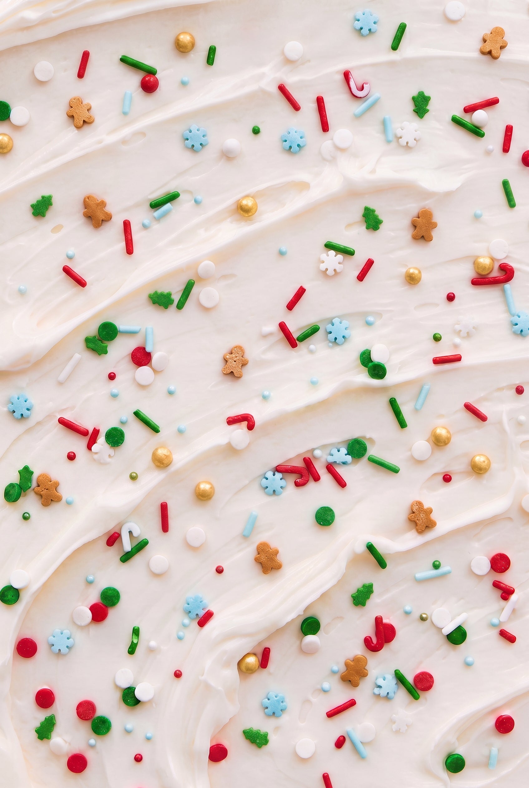 A close-up of white whipped frosting or cream swirled with colorful holiday sprinkles, including red, green, blue, gold, and brown shapes.